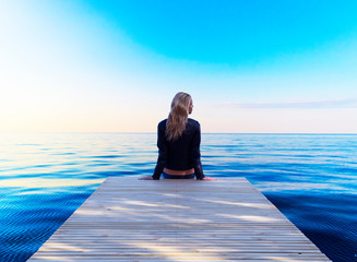 A lonely young blonde woman sitting wooden pier. Girl sitting on a wooden pier and looking at the...