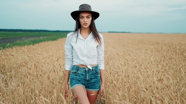 Young girl in shorts and hat walking down the wheat fields, smiling in the camera and touching the grains. Sun flare shines in the background. Nature is beautiful. Modern farming, happy youth and