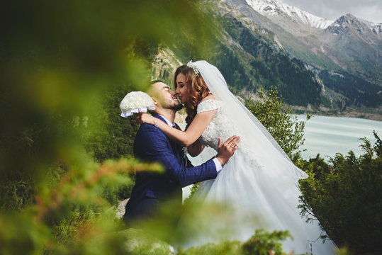 Beautiful wedding photo on mountain lake. Happy Asian couple in love, bride in white dress and groom in suit are photographed against background of the Kazakh landscape