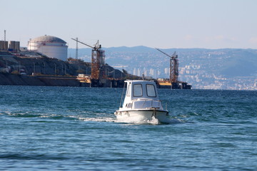 Local small white fishing boat coming in local harbour surrounded with blue sea and industrial complex in background on warm sunny summer day