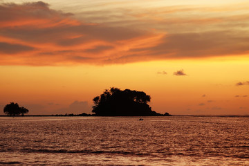 Sunset on a beach, silhouettes of tropical islands with palm trees in dark sea. Dramatic orange sky with clouds, romantic vacation concept