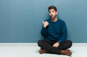 young handsome man sitting on floor looking astonished in disbelief, pointing at object on the side...