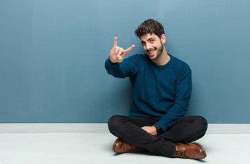 young handsome man sitting on floor feeling happy, fun, confident, positive and rebellious, making...