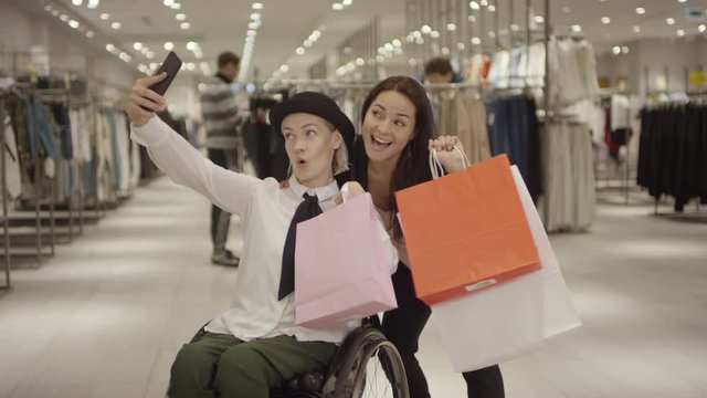 Tracking of happy beautiful woman in wheelchair holding shopping bag and taking selfie with her laughing female friend in clothing store