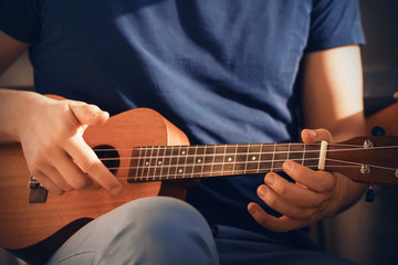 Fototapeta premium A man in a dark blue t-shirt and blue jeans plays a cheerful tune on a ukulele, sitting in the sunlight.