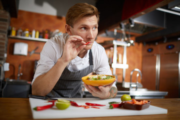 Portrait of handsome professional chef tasting red chili pepper while cooking Asian dish in...