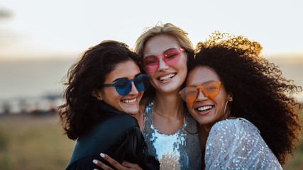 Three happy women wearing sunglasses hugging at evening outdoors