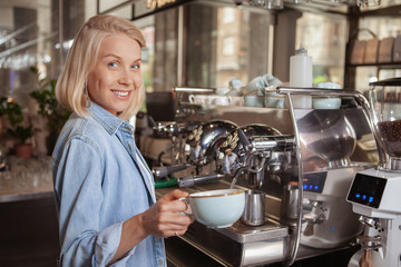 Beautiful happy female barista holding a cup of delicious aromatic coffee, smiling to the camera, copy space. Charming woman working at her coffee shop, preparing hot drinks for customers
