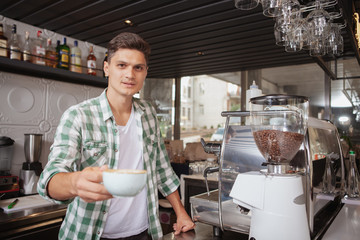 Charming male barista smiling, holding out a cup of delicious hot coffee to the camera, copy space. Handsome young man working at the coffee shop. Occupation, job concept