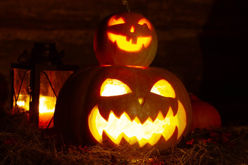 two halloween pumpkins in dark: one pumpkin stands on top of another, carved out eyes and terrible smiles glowing in the dark, lantern on wooden background