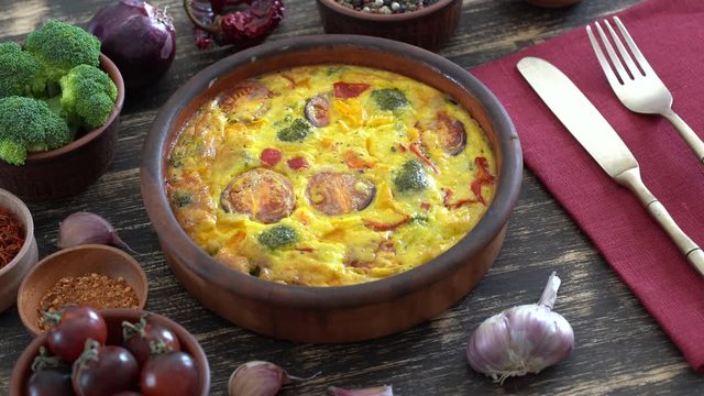 Ceramic bowl with vegetable frittata, simple vegetarian food. Frittata with egg, tomato, pepper, onion, broccoli and cheese on wooden table, close up. Italian egg omelette, rotates