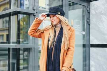 Fashionable beautiful long hair blonde girl in brown coat and blue cap at city, street style