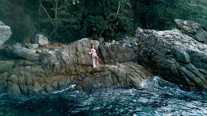 Aerial view: Sexy photo of a beautiful blonde posing standing in a half-naked white dress among the rocks near the sea shore with waves. Drone photo.