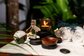 A lit candle and a jar of aromatic oils standing on stones for stone therapy and located on a terry towel next to which are transparent spheres, a  towel of white color and a sprig of lavender