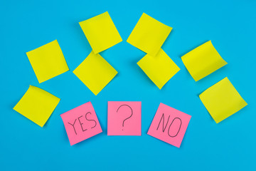 Fototapeta na wymiar Photo two pink stickers with the phrase yes or no and a sticker with a question mark on a blue background with yellow self-adhesive stickers. Concept of office stationery.