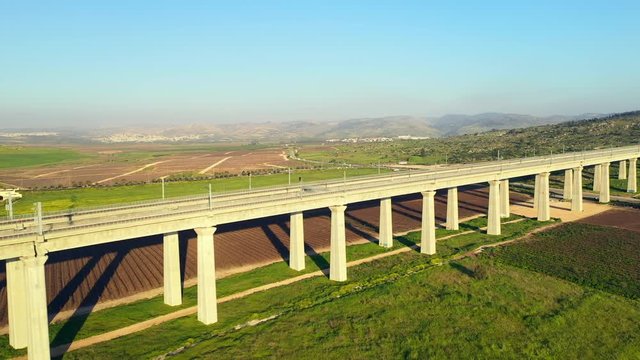 Aerial: A Rail Overpass Cuts Over Lush Fields and Farmland on Sunny Day Near Modiin