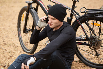 Close up portrait of cyclist resting in open air, sitting on ground in field or forest and talking on phone, wears sport wear and cap, having serious conversation with friend or wife while rides bike.