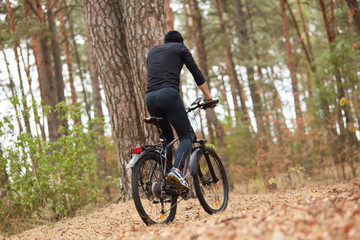 Fototapeta na wymiar Handsome young man biking in countryside, guy wearing black sportwear and cap posing backwards around trees in wood, spending spare time in open air, enjoys beautiful nature. Sport and fitness concept