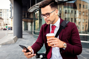 Serious young businessman with electric scooter standing in front of modern business building looking at phone.