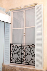Old mediterranean window. French doors with wrought-iron balconies. A tiny balcony with wrought iron railings against a cream stone building with open shutters and closed French doors. Home interior