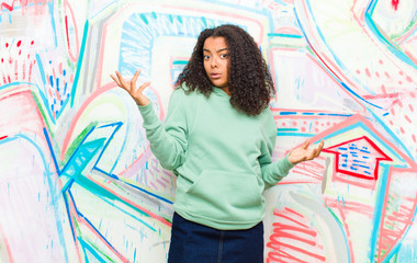 young pretty african american woman shrugging with a dumb, crazy, confused, puzzled expression,...