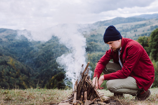 Outdoor horizontal image of young man in red jacket, knit beanie prepare brunches, for bonfire in mountains. Traveler man with beard, sitting near to campfire. Explore travel real wilderness lifestyle
