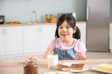 Funny little girl eating tasty toasts with chocolate spreading in kitchen