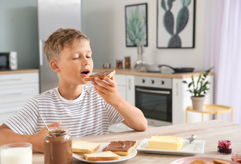 Funny little boy eating tasty toasts with chocolate spreading in kitchen