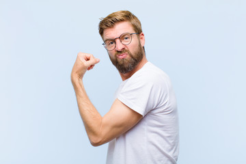 young blonde adult man feeling happy, satisfied and powerful, flexing fit and muscular biceps, looking strong after the gym