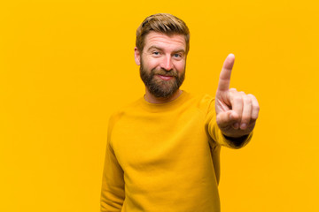 young blonde man smiling and looking friendly, showing number one or first with hand forward, counting down against orange wall © kues1