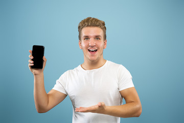 Caucasian young man's half-length portrait on blue studio background. Beautiful male model in shirt. Concept of human emotions, facial expression, sales, ad. Showing phone screen, payment, betting.