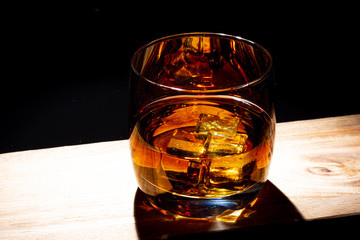 Glass of whiskey with ice on a wooden board. Wooden saw cut. Black background. Whiskey / Brandy / Cognac / Rum. Glass with a strong drink.
