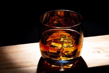 Glass of whiskey with ice on a wooden board. Wooden saw cut. Black background. Whiskey / Brandy / Cognac / Rum. Glass with a strong drink.