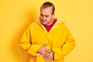 Young man wearing rain coat standing over isolated yellow background with hand on stomach because indigestion, painful illness feeling unwell. Ache concept.