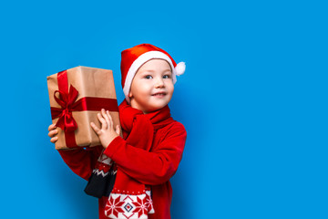 little boy in Santa's hat with cozy gift box and red tape want to know what inside the box, put to his ear