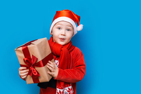 Christmas time. boy with Santa Claus Hat holding christmas gift with red tape and wery surprised face with open mouth
