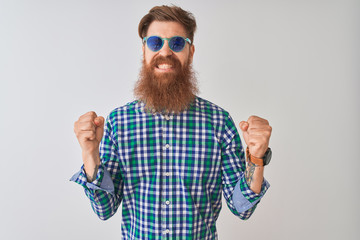 Young redhead irish man wearing casual shirt and sunglasses over isolated white background excited for success with arms raised and eyes closed celebrating victory smiling. Winner concept.