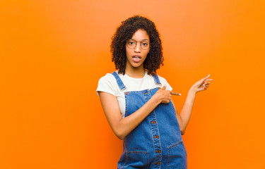 young pretty black woman feeling shocked and surprised, pointing to copy space on the side with...