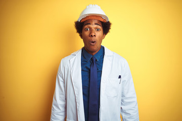Afro american engineer man wearing white coat and helmet over isolated yellow background afraid and shocked with surprise expression, fear and excited face.