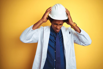 Afro american engineer man wearing white coat and helmet over isolated yellow background suffering from headache desperate and stressed because pain and migraine. Hands on head.