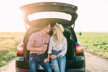Young happy couple sitting in car trunk outdoor. Road trip - obrazy, fototapety, plakaty