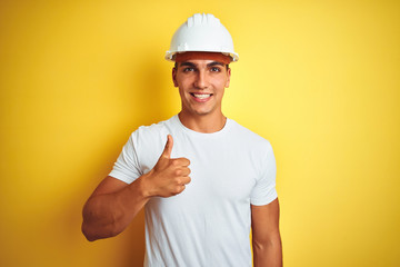 Young handsome man wearing construction helmet over yellow isolated background doing happy thumbs...
