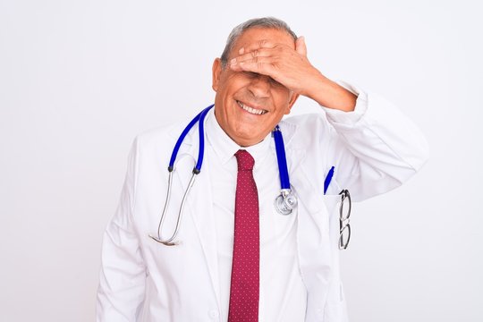 Senior grey-haired doctor man wearing stethoscope standing over isolated white background smiling and laughing with hand on face covering eyes for surprise. Blind concept.