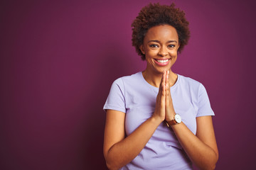 Young beautiful african american woman with afro hair over isolated purple background praying with...