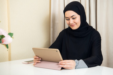 Muslim woman sitting at home with tablet computer, wearing earphones. Rest at home, preparing to watch a film or listen a music.