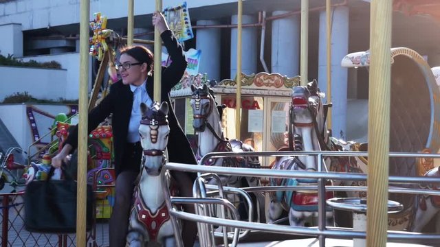 Funny scene. Young business woman riding a horse carousel. Girl throws up a briefcase while enjoying riding. Office workers entertainment concept. Slow motion 4k