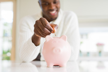 Handsome young african man putting a coin inside piggy bank smiling, excited about investment