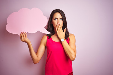 Beautiful woman holding speech bubble over pink isolated background cover mouth with hand shocked...