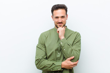 young handsome man looking happy and smiling with hand on chin, wondering or asking a question,...