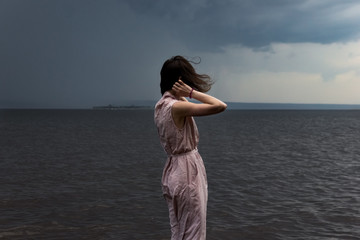 Fototapeta premium A cute young girl in a pink dress is standing in the water with flying hair during a storm and bad weather. Epic photo. Back view